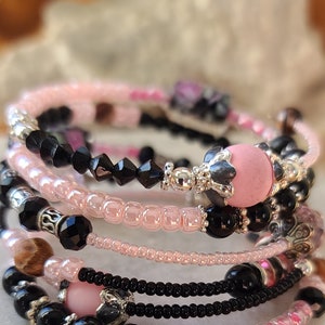 May include: Stack of beaded bracelets in shades of pink, black, and silver. The bracelets feature a variety of beads, including small pink seed beads, black faceted beads, and larger pink and silver accent beads. The bracelets are stacked together.