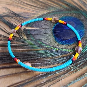 May include: A beaded anklet featuring turquoise, red, yellow, and brown beads. The anklet is displayed on a peacock feather. The beads are small and round, and the colours are vibrant. The anklet is a piece of jewellery.