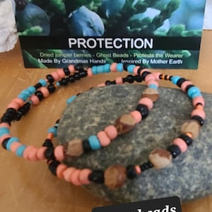 Könnte beinhalten: Zwei Perlenarmbänder aus getrockneten Wacholderbeeren, schwarzen Perlen und türkisfarbenen Perlen. Die Armbänder liegen auf einem Stein mit einem Schild, auf dem steht "Ghost Beads Protection - Dried juniper berries - Ghost Beads - Protects the Wearer Made By Grandmas Hands Inspired By Mother Earth".