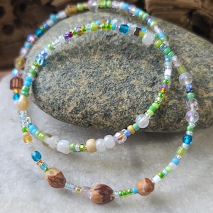 May include: Two beaded bracelets featuring a variety of small glass beads in shades of blue, green, white, and yellow, interspersed with clear faceted beads and three smooth, brown, organic-looking seed-like beads on the lower strand.