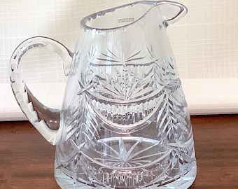 May include: A clear glass pitcher with a cut glass design. The pitcher has a wide mouth and a handle. The pitcher is made of crystal and has a delicate, intricate design.
