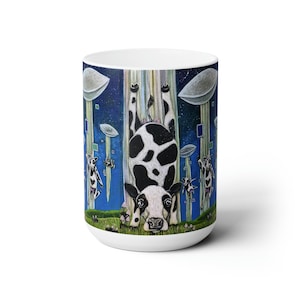 Pode incluir: Uma caneca de cerâmica branca com uma ilustração colorida de uma vaca preta e branca pendurada de cabeça para baixo em um campo de grama verde. A vaca está cercada por nuvens azuis e brancas e outras vacas.