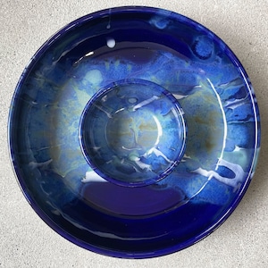 May include: A two-piece ceramic serving set with a deep blue glaze. The set includes a large, shallow dish and a smaller, round bowl nested inside. The glaze features streaks of lighter blue, white, and gold, creating a marbled effect.