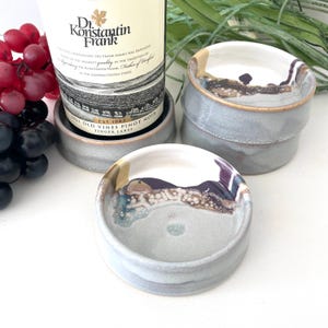 May include: Set of four gray ceramic coasters with a white glaze and brown, purple, and blue accents. The coasters are stacked on top of each other.