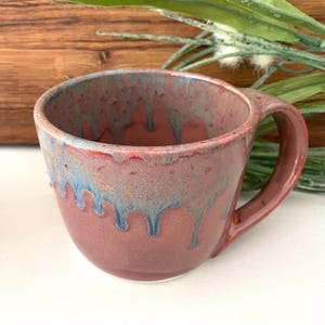 May include: A handmade ceramic mug with a rounded shape and a large handle. The mug features a blend of colors, including a base of muted red-brown with blue and gold drips around the rim. The mug is suitable for coffee or tea.