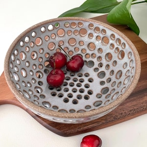 May include: A gray ceramic bowl with a perforated design, filled with ripe red cherries. The bowl has a textured surface and a brown rim. The bowl is placed on a wooden cutting board.