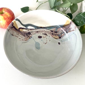 May include: A large, white ceramic bowl with a light gray glaze. The bowl has a unique, hand-painted design with brown, blue, and gold accents.
