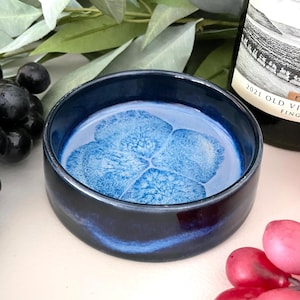 May include: A small, round, dark blue ceramic bowl with a light blue floral design in the center. The bowl has a glossy finish and is surrounded by grapes and greenery. A wine bottle is partially visible in the background.