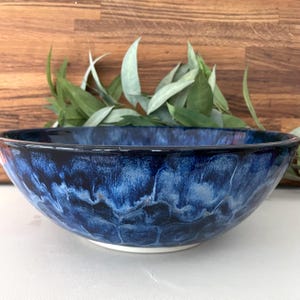 May include: A large, handmade ceramic bowl with a deep blue glaze. The bowl features a white base and a dark blue rim. The interior has a marbled effect with lighter blue hues. The bowl is suitable for serving salads or as a decorative piece.