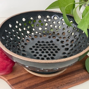 May include: A black ceramic colander bowl with numerous circular perforations. The bowl has a wide rim with a natural, unglazed edge. The colander sits on a matching base, ideal for draining fruits or vegetables. The bowl is approximately 10 inches in diameter.