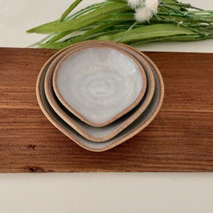 May include: A set of three nesting ceramic dishes with a light blue glaze and a natural brown rim. The dishes are heart-shaped and arranged on a wooden surface, with greenery in the background. The dishes are ideal for serving small portions.
