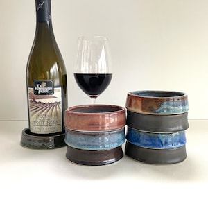 May include: A wine bottle and a glass of red wine are next to a stack of handmade ceramic bowls. The bowls are in shades of brown, blue, and gray. The wine bottle is labeled "Pinot Noir" from the Finger Lakes region.