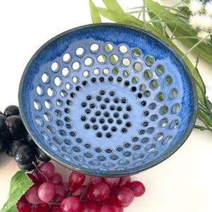 May include: A blue ceramic bowl with numerous circular perforations. The bowl has a gradient of blue hues, with a darker rim and a lighter center. The bowl is likely used for serving or displaying food items.