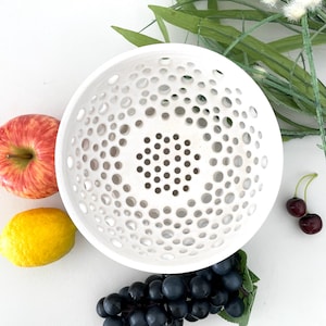 May include: White ceramic bowl with a circular pattern of holes. The bowl is perfect for holding fruit or vegetables.