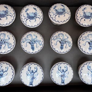 May include: A set of twelve white ceramic cabinet knobs with blue sea creature designs. The knobs feature crabs, octopuses, and lobsters, each with a scalloped edge. The knobs are arranged in a black box.