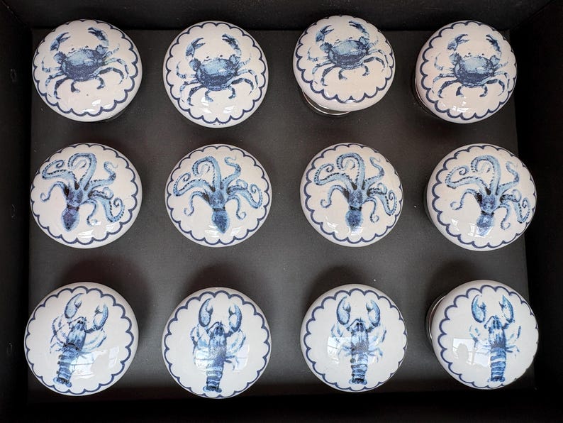 May include: A set of twelve white ceramic cabinet knobs with blue sea creature designs. The knobs feature crabs, octopuses, and lobsters, each with a scalloped edge. The knobs are arranged in a black box.