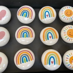 May include: Set of 10 ceramic knobs with various designs, including rainbows, hearts, and suns. The knobs are white with colorful designs.