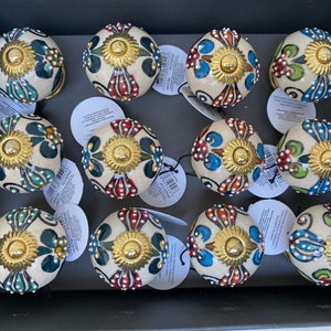 May include: Twelve ceramic knobs with a white base and colorful floral designs. Each knob has a gold center and a small, raised dot pattern around the edge.