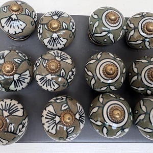 Könnte beinhalten: Eine Sammlung von zwölf Keramik-Schrankknöpfen. Die Knöpfe sind olivgrün mit weißen Blumenmustern und goldfarbener Hardware. Die Knöpfe sind auf einer dunkelgrauen Oberfläche angeordnet.