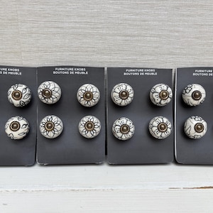 Può includere: Set di 20 pomelli per cassetti in ceramica bianca con motivi floreali neri e dettagli dorati. I pomelli sono esposti su quattro schede con il testo "Furniture Knobs Boutons de Meuble".