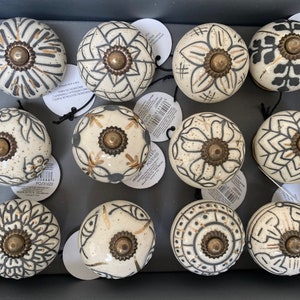 May include: A selection of ceramic cabinet knobs with cream, black, and gold patterns. Each knob features a brass-toned base. The designs include floral and geometric motifs. The knobs are displayed in a dark grey box.