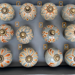 Op de afbeelding: Twaalf keramische knoppen met een grijze achtergrond en oranje bloemmotieven. Elke knop heeft een messingkleurig accent in het midden.