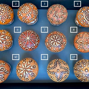 May include: Twelve ceramic knobs with intricate white designs on an orange background. The knobs are arranged in a grid pattern on a dark grey surface.