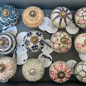 May include: Twelve ceramic knobs with various patterns and colors. The knobs are round and have a small metal knob in the center. The patterns include geometric shapes, flowers, and dots. The colors include white, black, blue, red, and yellow.