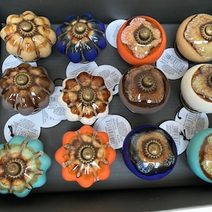 May include: Assortment of 12 ceramic cabinet knobs in various colours and patterns. The knobs feature a flower-shaped design with a brass accent in the centre.
