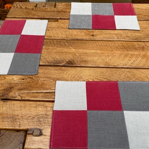 May include: Three placemats with a checkered pattern in shades of grey, white, and red. The placemats are made of a woven fabric and have a subtle texture.