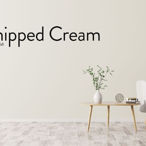 May include: A living room interior design with a light gray sofa, a wooden coffee table, a vase with green plants, and a white wall with the text "Whipped Cream Nordic Chic Wall".