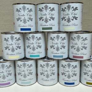 May include: A collection of twelve paint cans stacked in a pyramid shape. The cans are all white with black lettering and a floral design. The text on the cans reads "Nordic Chic Furniture Paint". The cans are in various shades of blue, green, purple, yellow, and brown.