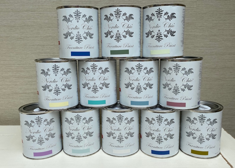 May include: A collection of twelve paint cans stacked in a pyramid shape. The cans are all white with black lettering and a floral design. The text on the cans reads "Nordic Chic Furniture Paint". The cans are in various shades of blue, green, purple, yellow, and brown.