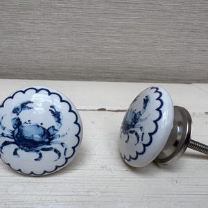 May include: Two white ceramic cabinet knobs with a blue crab design. The knobs have a scalloped edge and a silver metal base with a threaded screw for installation. The crab design is a detailed illustration.