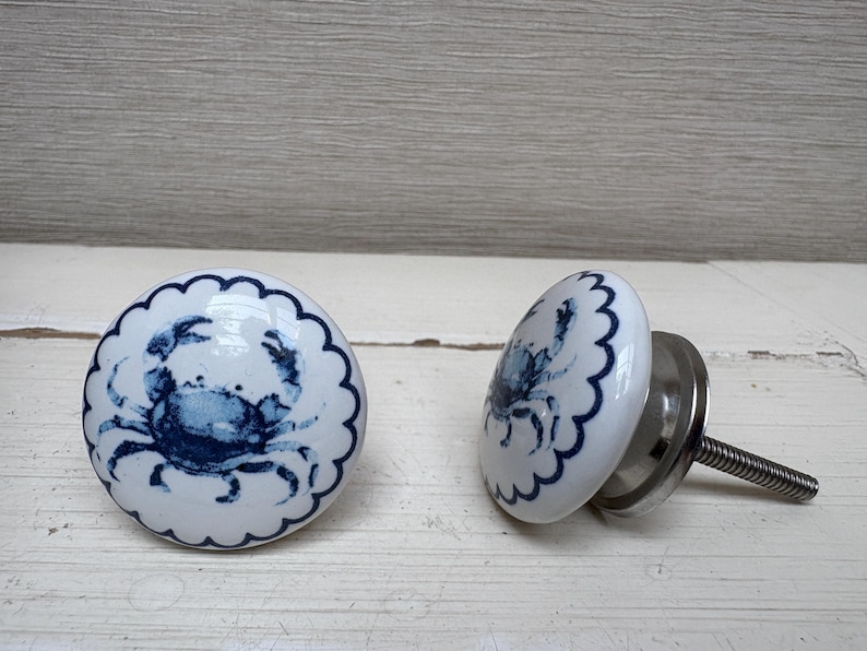 May include: Two white ceramic cabinet knobs with a blue crab design. The knobs have a scalloped edge and a silver metal base with a threaded screw for installation. The crab design is a detailed illustration.