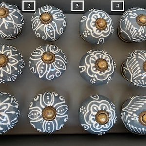 Op de afbeelding: Twaalf keramische ladegrepen met een grijze basis en witte bloemmotieven. Elke knop heeft een messing accent in het midden.