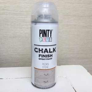 May include: A can of Pinty Plus Chalk Finish spray paint in the colour Piedra Stone. The can is white with a clear cap and features the brand name and product information. The label shows a painted dresser.