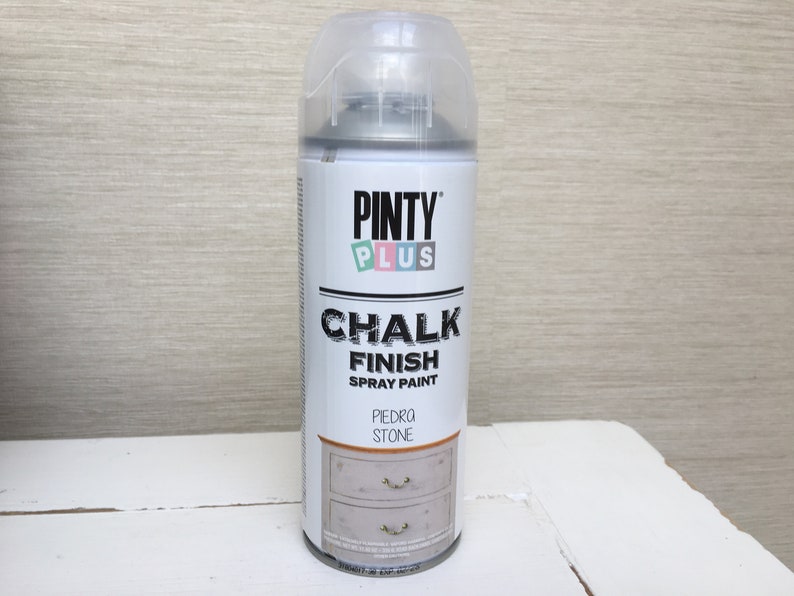May include: A can of Pinty Plus Chalk Finish spray paint in the colour Piedra Stone. The can is white with a clear cap and features the brand name and product information. The label shows a painted dresser.