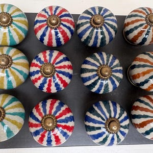 Pode incluir: Doze puxadores de armário de cerâmica com ferragens de latão. Os puxadores apresentam um design listrado em várias cores, incluindo azul, vermelho, amarelo e castanho. Os puxadores estão dispostos sobre uma superfície escura.