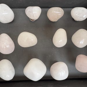 Könnte beinhalten: Eine Sammlung von 11 glatten, weißen und rosafarbenen Steinen, einige mit einem leichten Schimmer, auf einer schwarzen Oberfläche angeordnet.