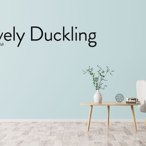 May include: A light blue wall with the text "Lovely Duckling" and "Nordic Chic Wall" in black lettering. A white couch with wooden legs is in the lower right corner of the image. A small wooden table with a vase of green plants and a decorative object is in the center of the image.