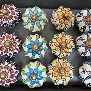 May include: A collection of twelve ceramic cabinet knobs with floral designs. The knobs feature a variety of colors, including red, blue, orange, and green, with brass-colored hardware. Each knob is approximately 2 inches in diameter.