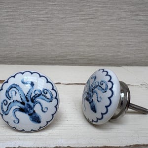 May include: Two white ceramic cabinet knobs with a blue octopus design. The knobs have a scalloped edge and a silver metal screw. The octopus design is centered on the knob.