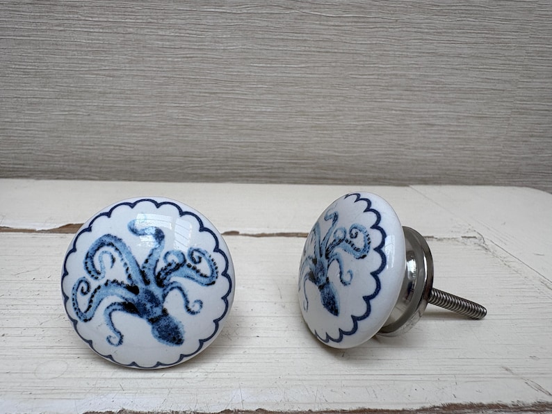 May include: Two white ceramic cabinet knobs with a blue octopus design. The knobs have a scalloped edge and a silver metal screw. The octopus design is centered on the knob.