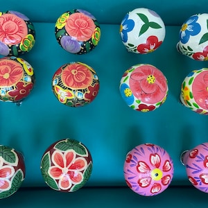 Pode incluir: Doze botões de madeira pintados à mão com desenhos florais em várias cores, incluindo vermelho, rosa, azul, verde e amarelo. Os botões estão dispostos em uma grade sobre um fundo turquesa.