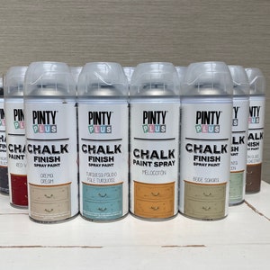 May include: A selection of Pinty Plus Chalk Finish spray paint cans. The cans are white with clear lids and display the "Pinty Plus" logo and colour names such as "Crema Cream" and "Beige Sahara". The cans are arranged on a white surface.