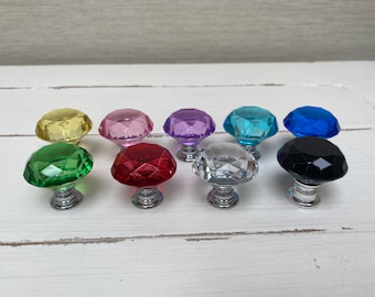 Puede incluir: Una colección de diez pomos de cristal facetado en varios colores, incluyendo amarillo, rosa, morado, turquesa, azul, verde, rojo, transparente y negro. Cada pomo tiene una base plateada.