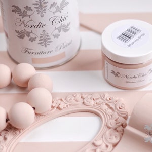 Puede incluir: Primer plano de una lata de pintura y un frasco etiquetados como "Nordic Chic Furniture Paint" en el color "Blushing Bride". La pintura es de color rosa claro y se muestra junto a un marco decorativo y cuentas de madera.