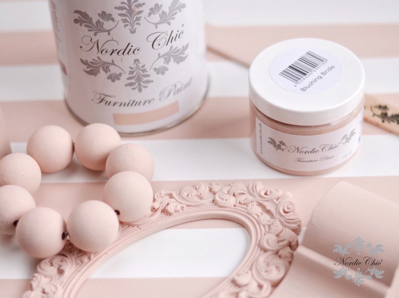Puede incluir: Primer plano de una lata de pintura y un frasco etiquetados como "Nordic Chic Furniture Paint" en el color "Blushing Bride". La pintura es de color rosa claro y se muestra junto a un marco decorativo y cuentas de madera.
