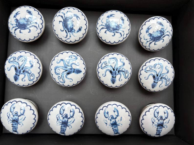 May include: A collection of twelve white ceramic cabinet knobs with blue marine life designs. The knobs feature crabs, octopuses, and lobsters, each with a scalloped edge. These decorative knobs are ideal for adding a nautical touch to furniture.
